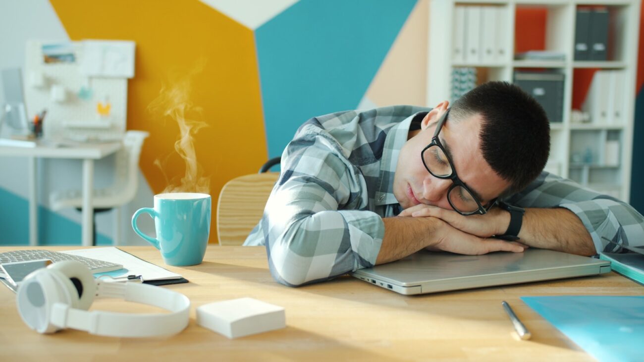 Man asleep at his desk with coffee and a laptop, illustrating feeling tired after eating lunch