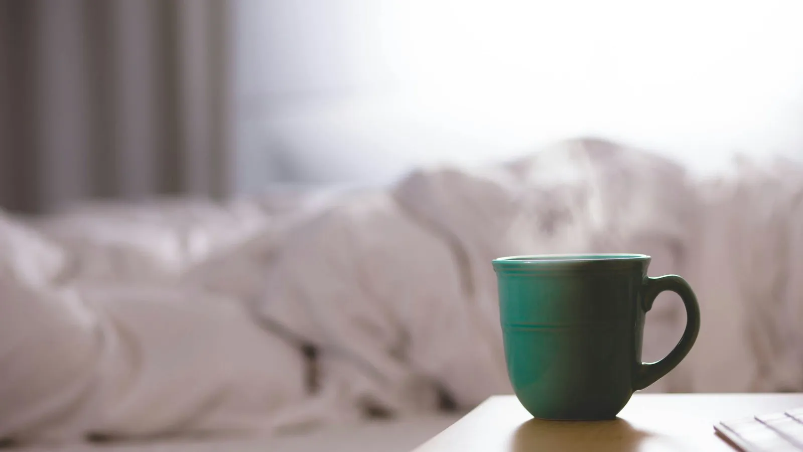 Caffeine crash: a green coffee mug on a bedside table beside an unmade bed in soft morning light